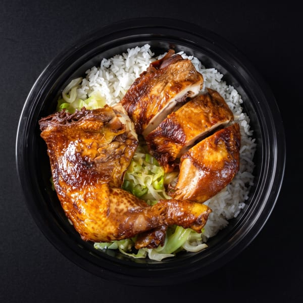 Grilled chicken pieces with rice and cabbage in a black takeout bowl on a dark surface.