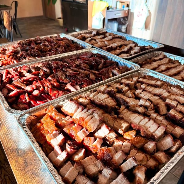 Multiple foil trays filled with sliced char siu, roasted pork belly, and other meats.