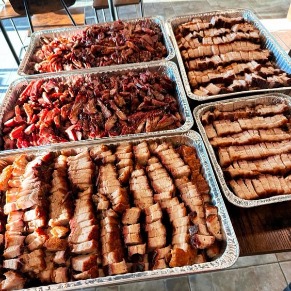 Multiple foil trays filled with sliced roasted pork belly and char siu on a wooden table.